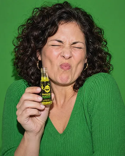 Frau mit dunklen Locken hält ein offenes Fläschchen und spitzt sauer die Lippen